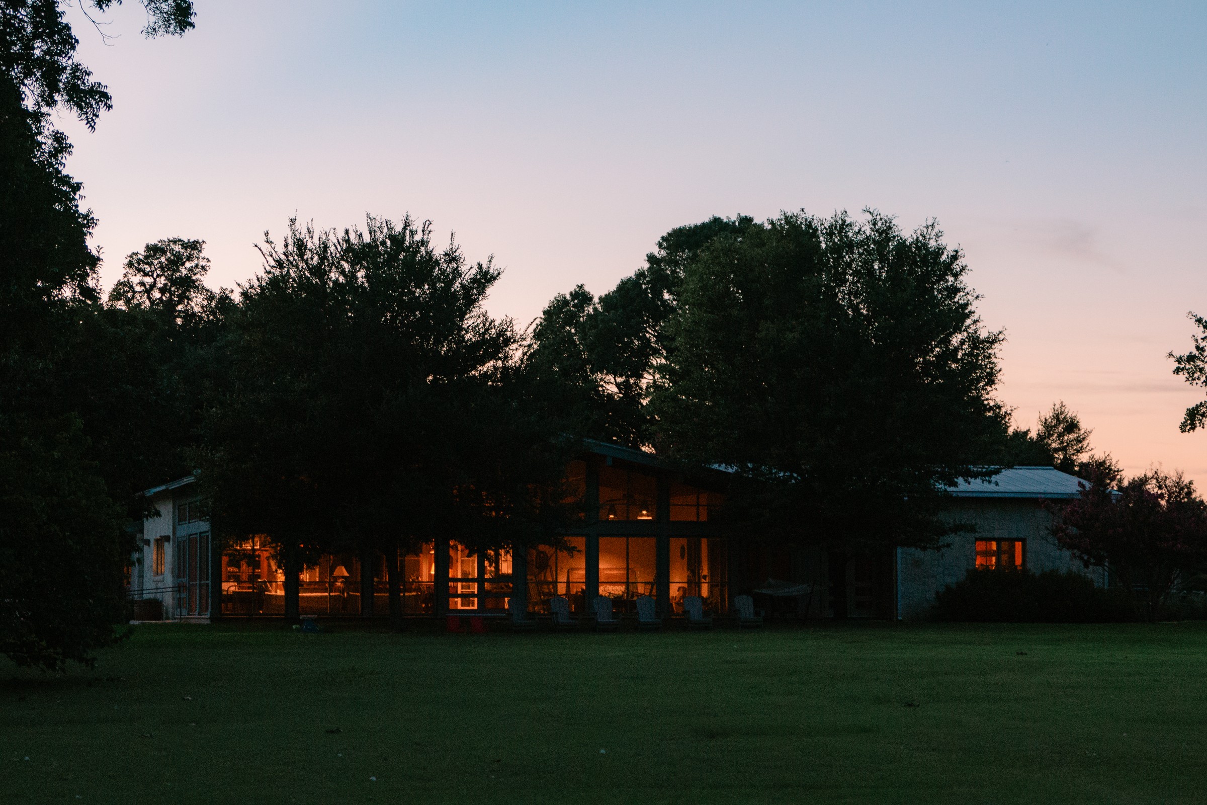 A home glowing at dusk, warm light spilling from its windows across a wooded lawn